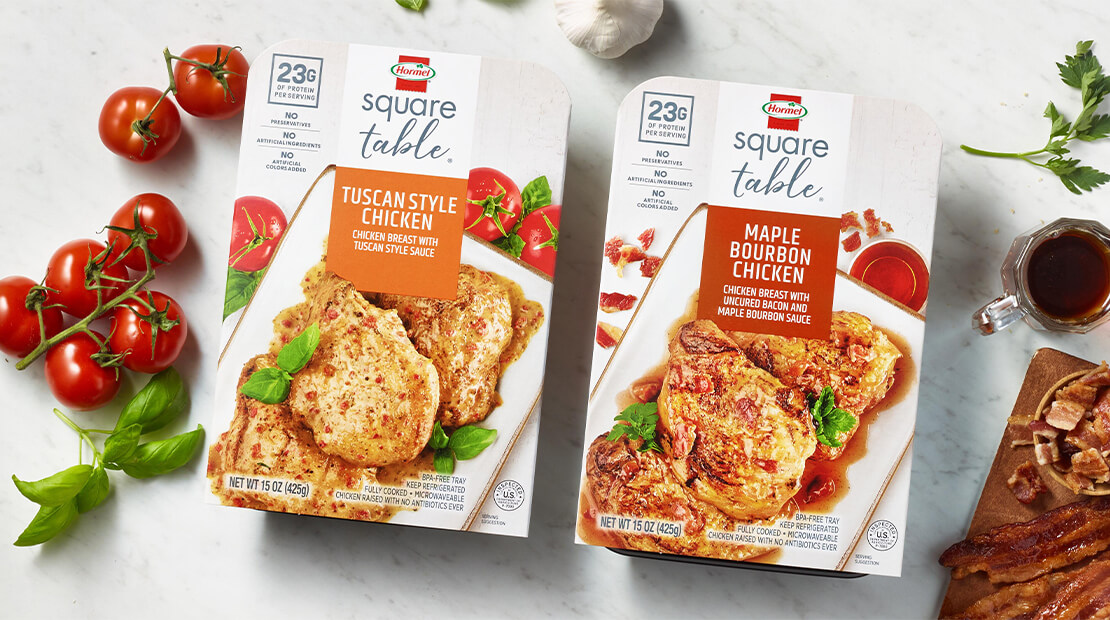 HORMEL® SQUARE TABLE® Tuscan Style Chicken and HORMEL® SQUARE TABLE® Maple Bourbon Chicken on a white marble counter top with tomatoes and a small dish of maple syrup next to the packages.